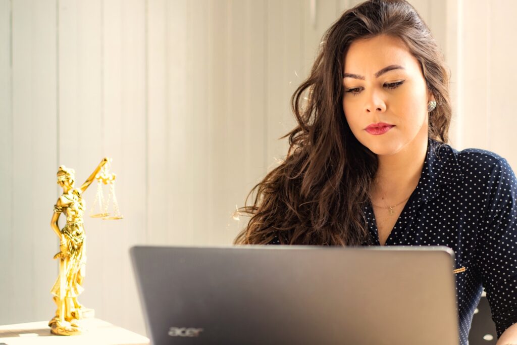 Na zdjęciu znajduje się kobieta patrząca w komputer, a obok na biurku stoi figurka Temidy